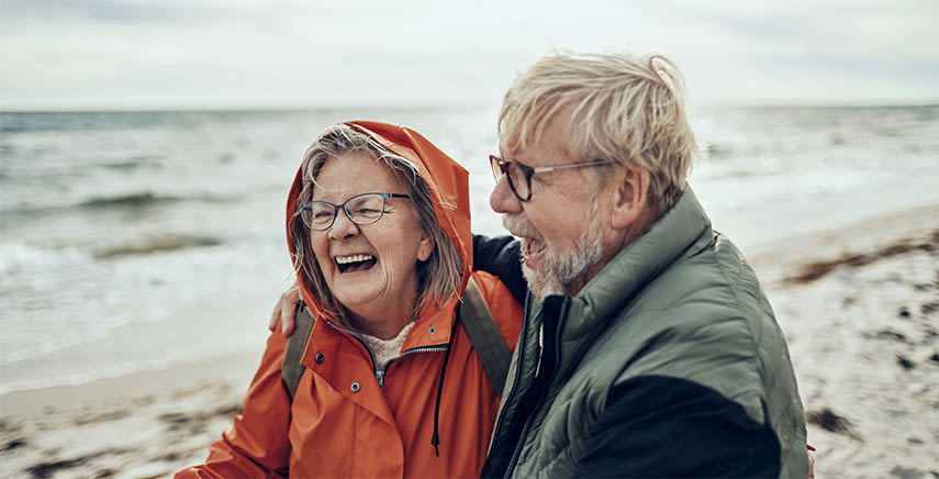 Ett par pensionärer står och håller om varandra på stranden och skrattar gott.