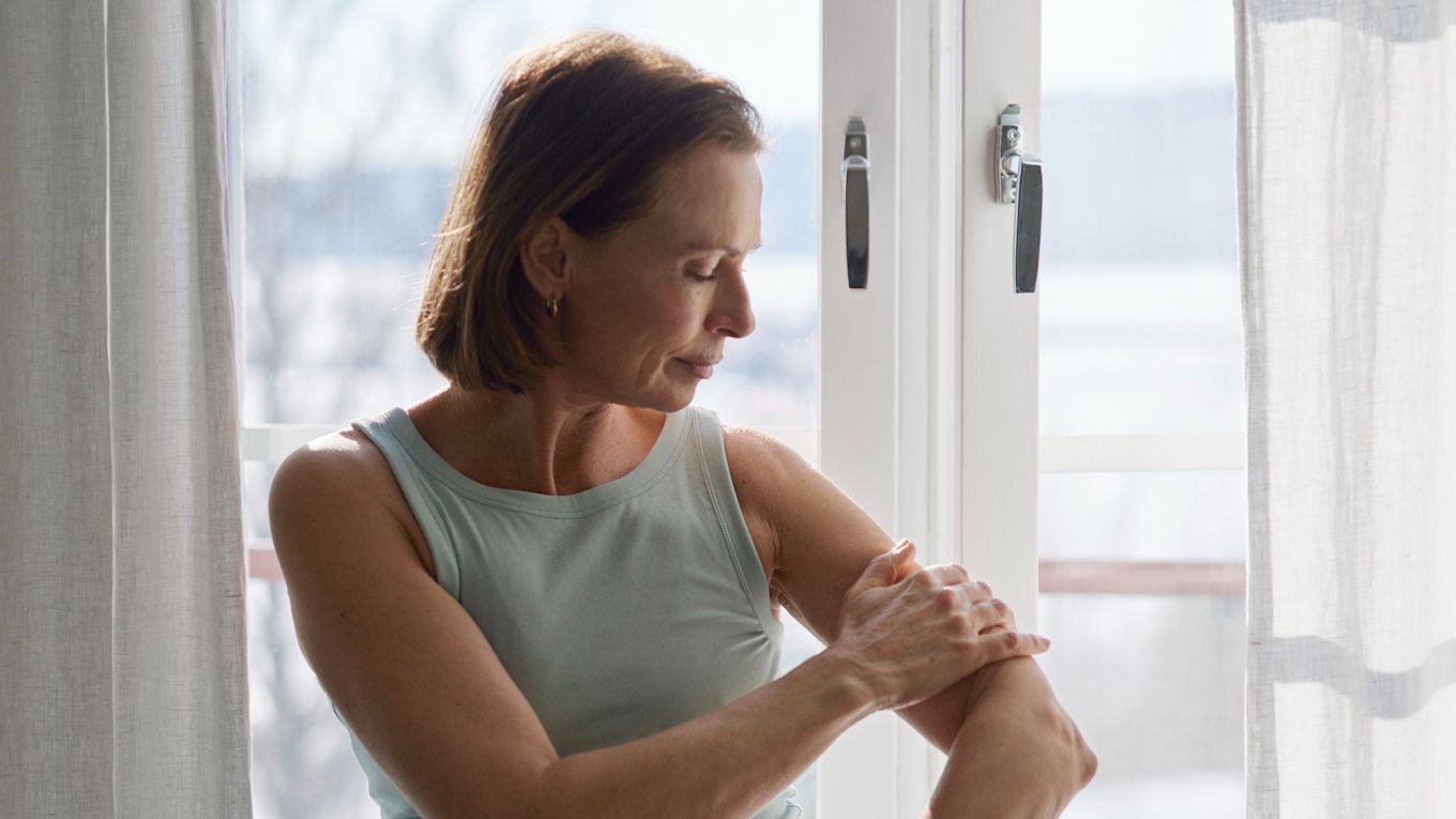 Kvinna som står framför ett fönster och applicerar hudkräm på sin arm. Hon är klädd i en ljusgrön linne omgiven av naturligt ljus som strömmar in genom fönstret. 