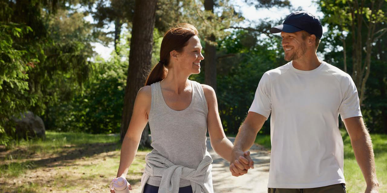 En kvinna och en man är ute och promenerar hand i hand i solen. Båda är glada och tittar på varandra.