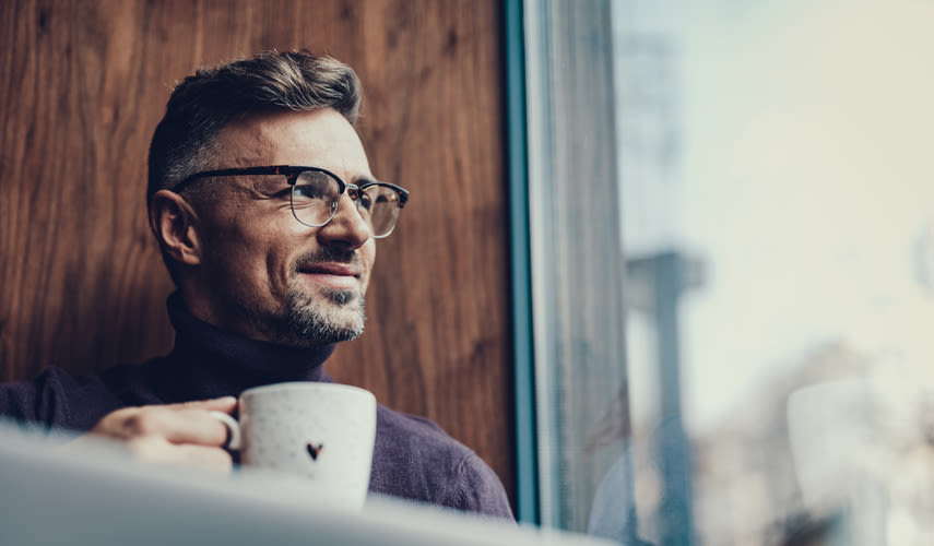 En man sitter och tittar ut genom fönstret och ler, men en kaffekopp i handen.