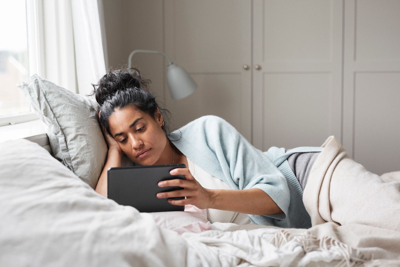 Kvinna ligger i sängen bland täcken och kuddar och tittar på en surfplatta.
