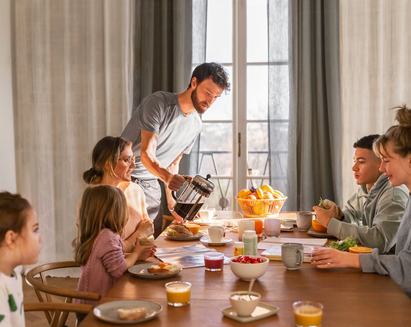 En stor familj sitter vid frukostbordet och äter tillsammans. På bordet finns smörgåsar, juice, kaffe, smoothies, frukt och yoghurt.