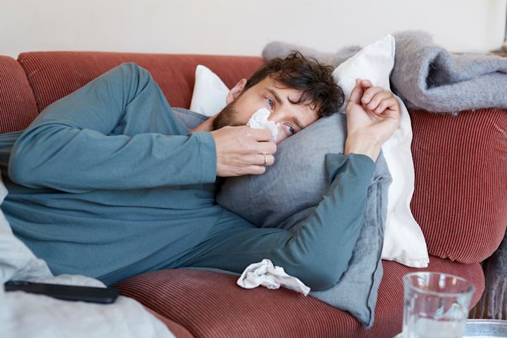 Man ligger i soffan och tittar på tv och snyter sig, med kuddar och filtar runtomkring sig.