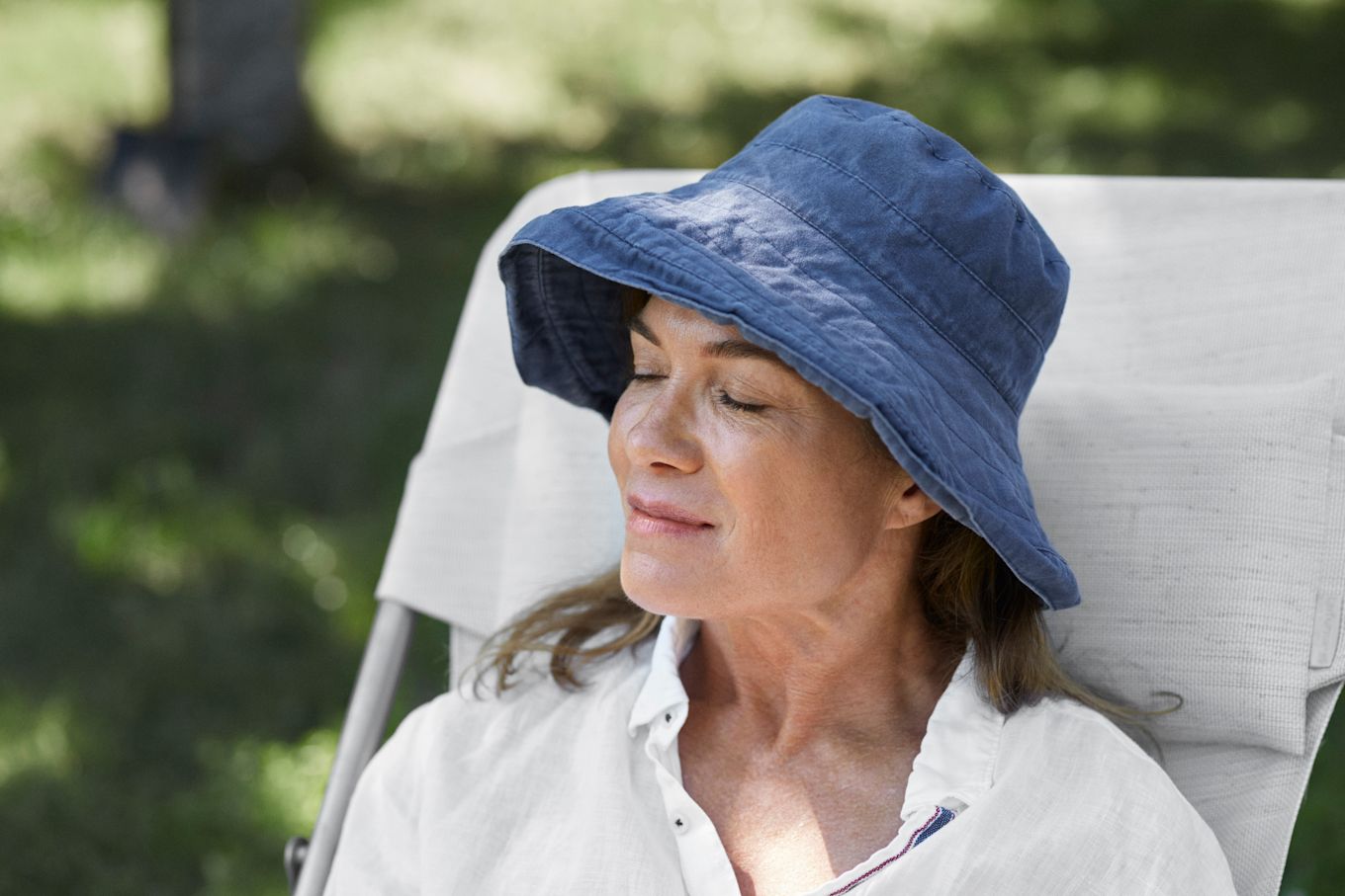 En kvinna i blå solhatt sitter och solar i naturen med slutna ögon.