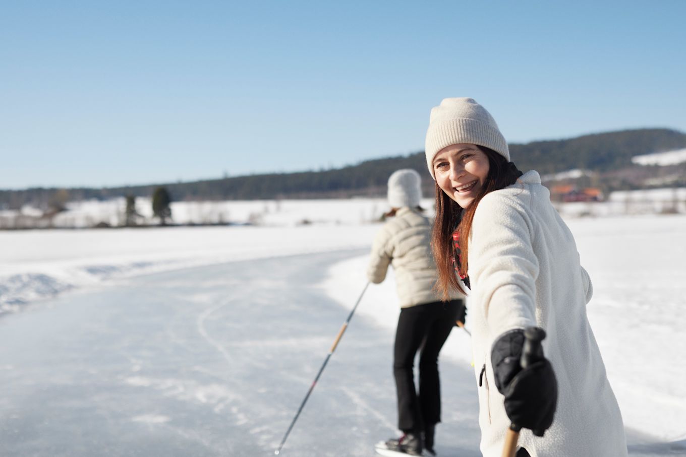 Två kvinnor är ute på isen under vintern och åker skridskor, den ena tittar tillbaka och ler.
