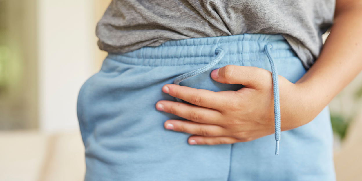 Kvinna som håller om sin mage, ansiktet syns inte. Hon bär blåa mjuka byxor och en grå t-shirt.