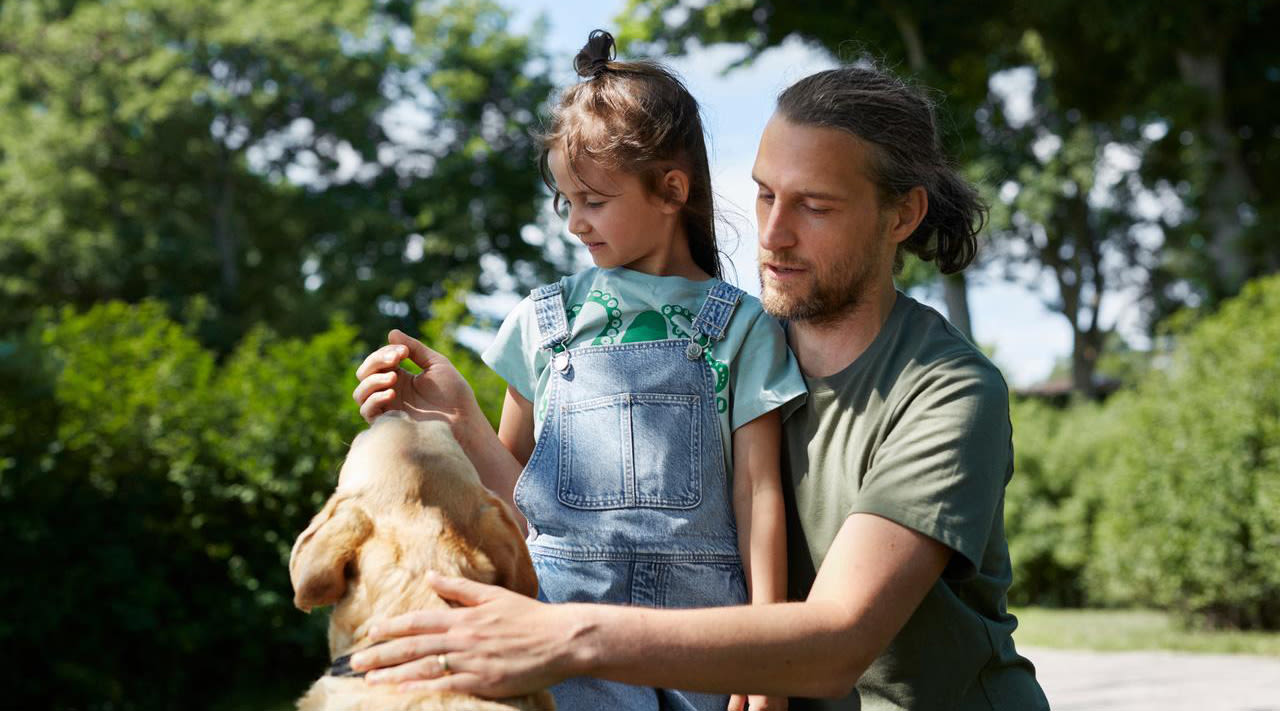 Pappa står på knä intill sin dotter och båda klappar en hund ute i naturen.