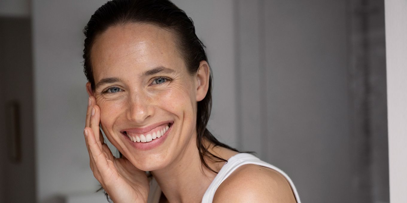 Kvinna tittar in i kameran och ler med ett stort leende. Hon håller några fingrar lätt på ena kinden.