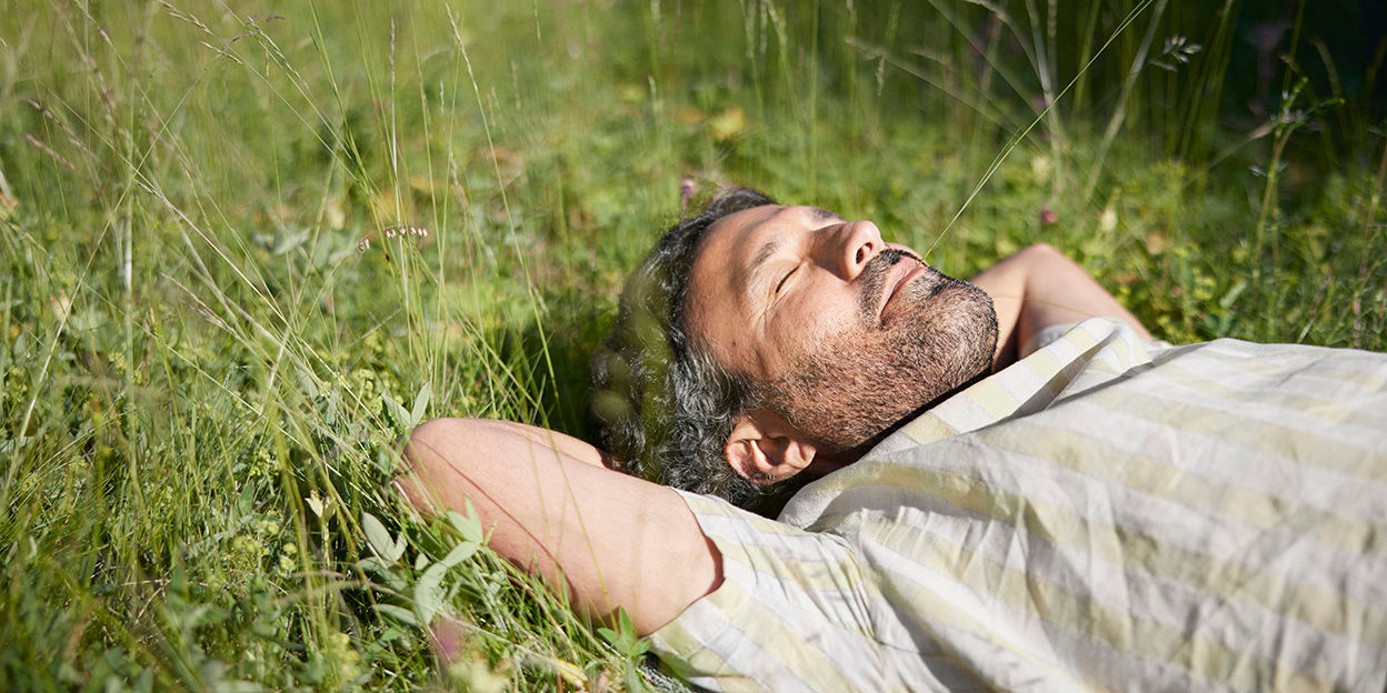 Man i gul och vitrandig skjorta ligger med stängda ögon på en sommaräng med ett grässtrå i mungipan och njuter av solskenet. 