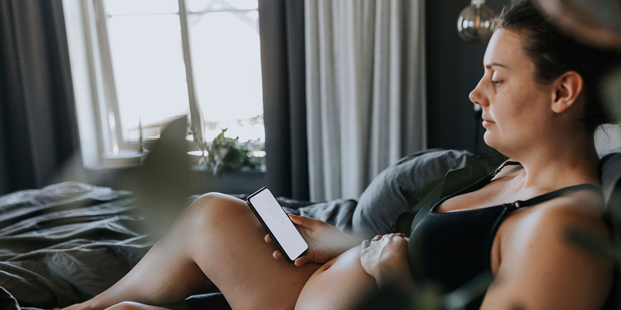 Gravid kvinna i underkläder sitter i sängen och tittar på sin telefon med en hand på magen.