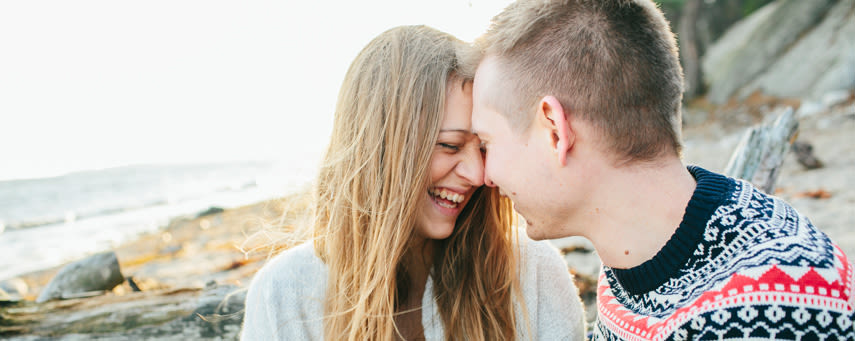 En ung tjej och kille sitter på en strand och skrattar med sina ansikten tätt ihop.