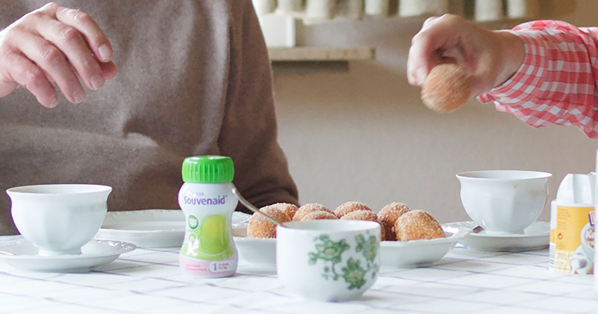 På ett matbord står en flaska Souvenaid. Två personer sitter intill bordet och sträcker sig framåt. På bordet står även kaffekoppar, en sockerskål, assietter och ett fat med fika.