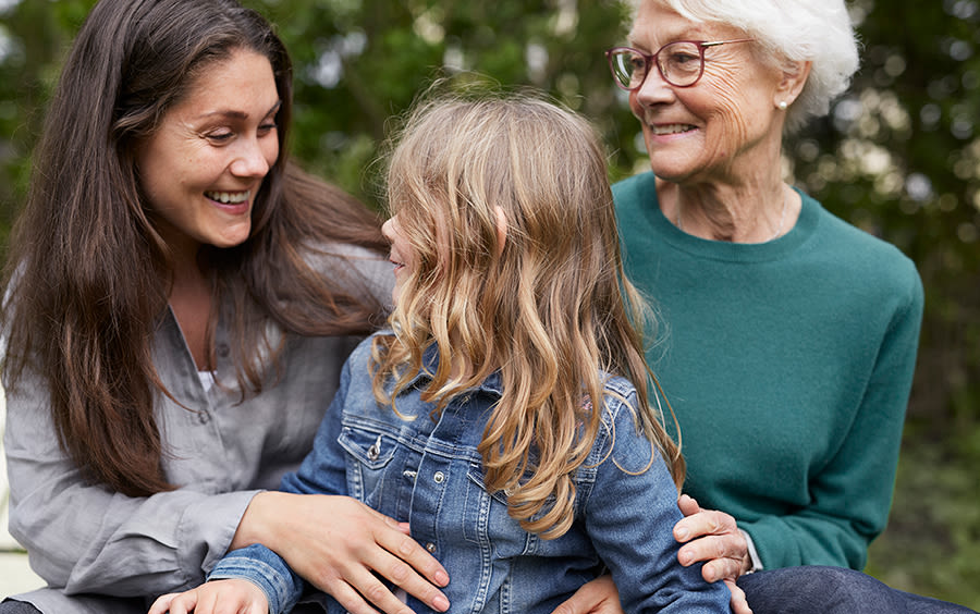 Ett barn sitter mellan sin mamma och mormor. Alla tre ser glada ut och håller om varandra.